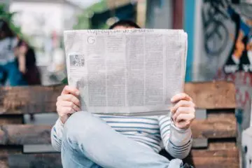 man in jeans sitting at a bench and reading a newspaper