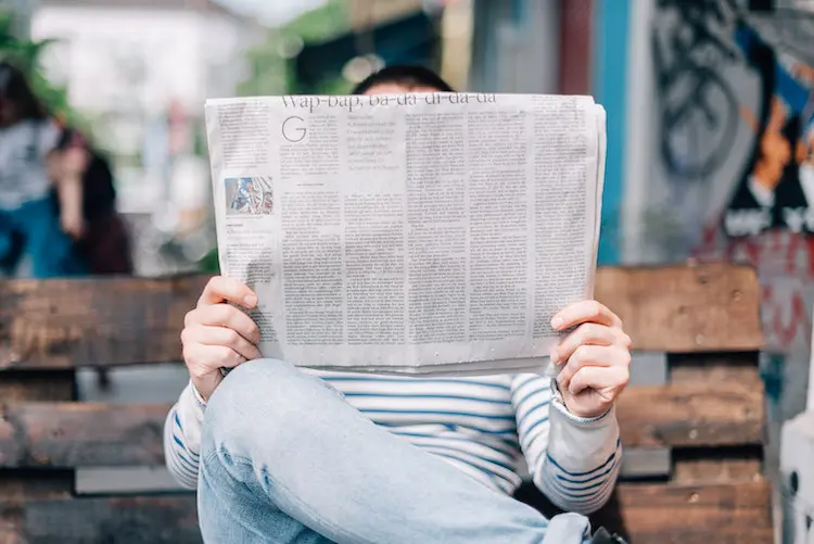 man with jeans sitting on a bench and reading a newspaper