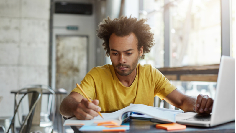 man in yellow shirt taking notes while studying with his laptop